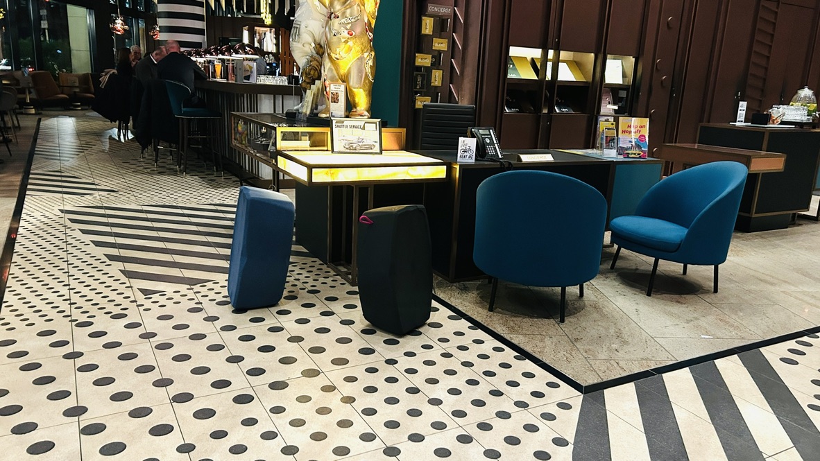 Innenansicht einer modernen Hotel-Lobby oder Lounge. Der Raum zeigt eine Rezeption/Counter mit einem Bodenmuster aus Schwarz-Weiß-Streifen und gepunkteten Fliesen. Im Vordergrund stehen zwei türkisfarbene, schwere Loungesessel neben zwei leichten, zylindrischen Hockblocs in Dunkelblau und Schwarz. Der Kontrast zwischen den festen, schweren und den flexiblen, mobilen Sitzgelegenheiten wird hervorgehoben.