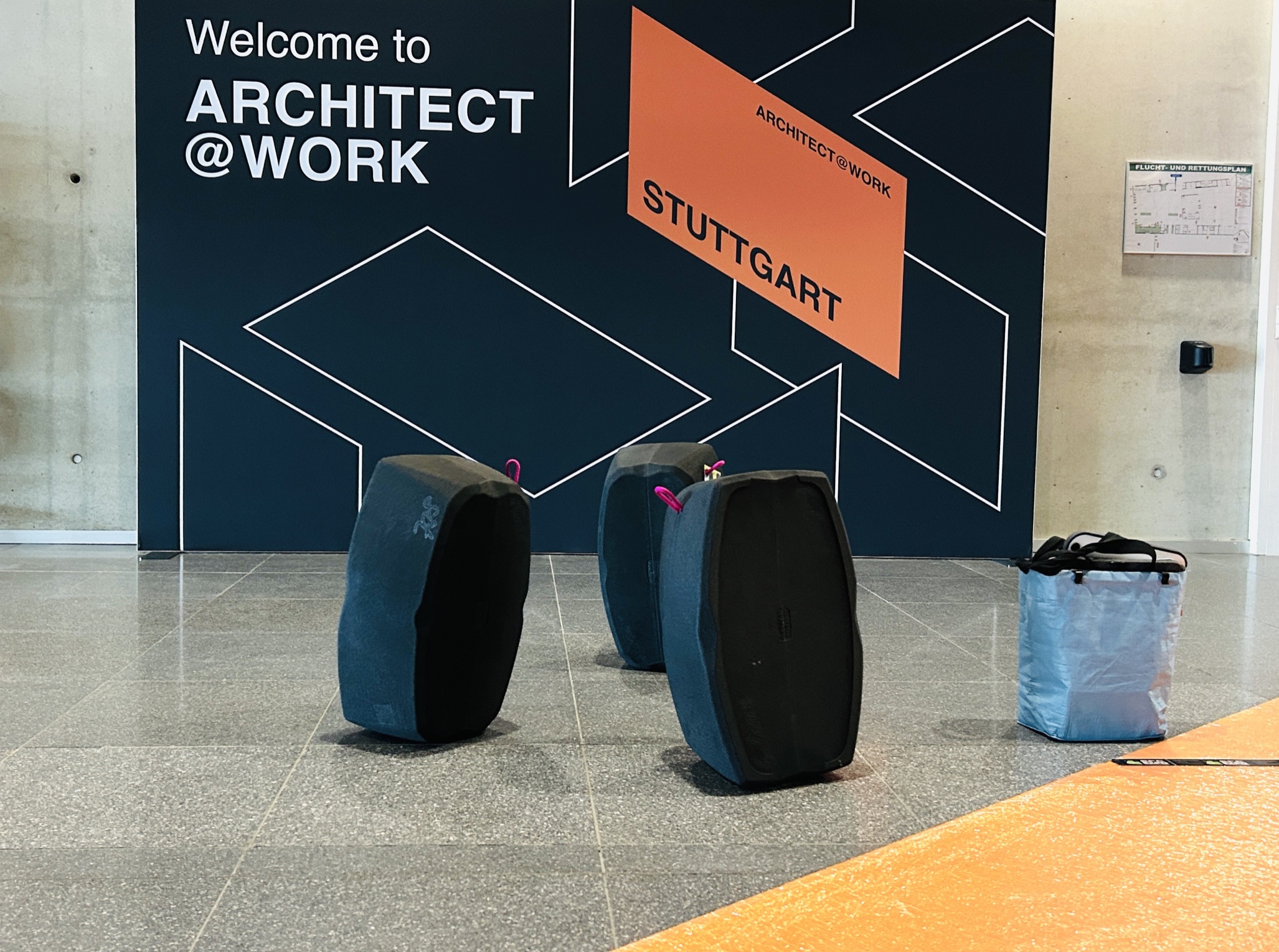 Eingangsbereich der Messe ARCHITECT@WORK STUTTGART. Im Vordergrund stehen drei dunkelblaue, ovale, mobile Sitzelemente (Hockblocs oder ähnliche ultraleichte Möbel) sowie eine hellblaue, rechteckige, taschenartige Transporteinheit der imsinne GmbH oder Wooden Valley gGmbH. Im Hintergrund ist auf einer dunkelblauen Wand in weißer und oranger Schrift das Logo "Welcome to ARCHITECT@WORK STUTTGART" zu sehen.