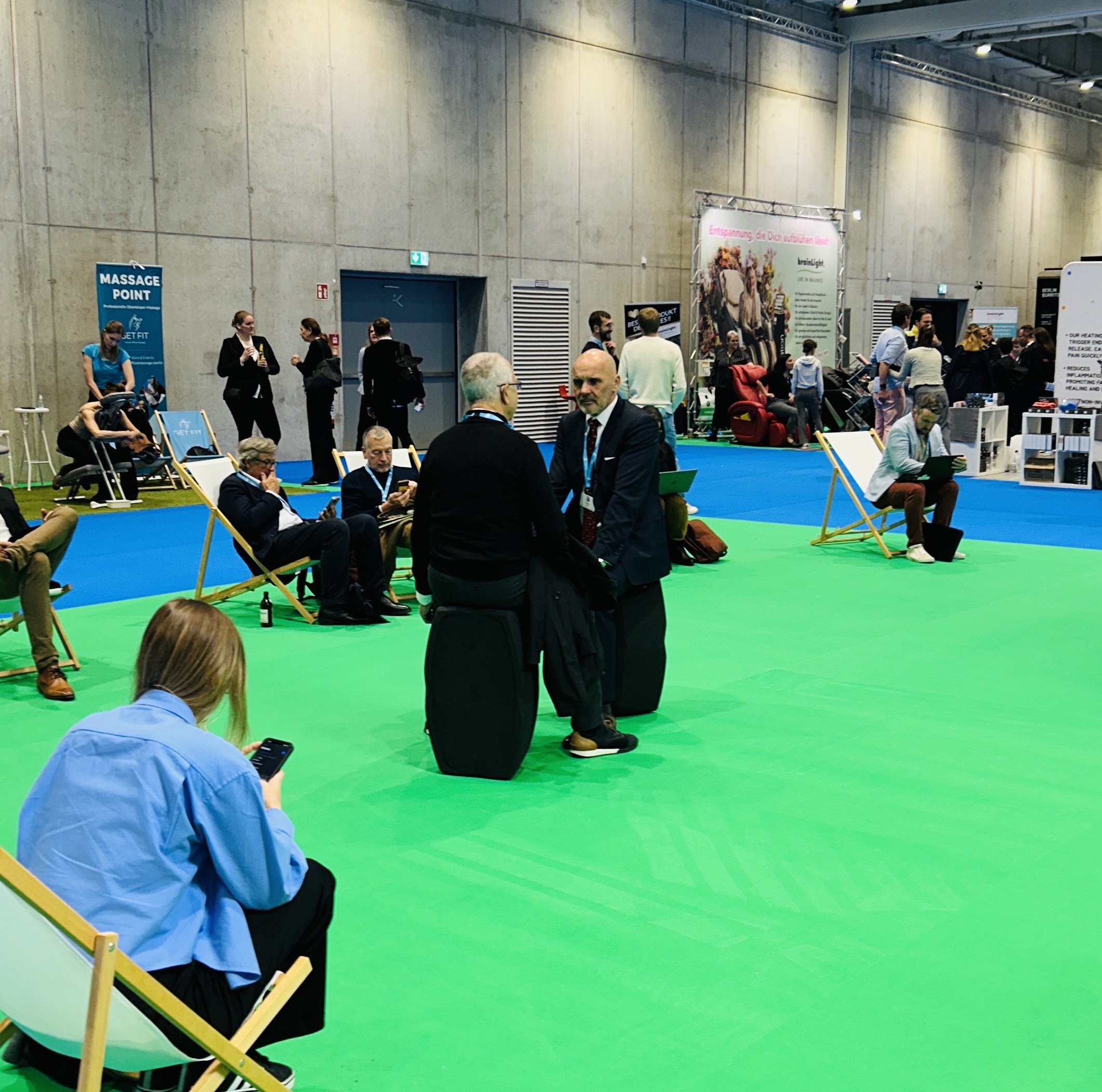 Innenansicht einer Messehalle mit einer Entspannungszone auf leuchtend grünem und blauem Teppich. Im Vordergrund sitzen zwei Geschäftsleute in Anzügen auf runden, schwarzen Hockblöcken und führen ein intensives Gespräch. Rundherum sitzen weitere Personen in Liegestühlen und nutzen ihre Mobiltelefone oder unterhalten sich. Links ist ein Schild mit der Aufschrift „MASSAGE POINT“ zu erkennen.