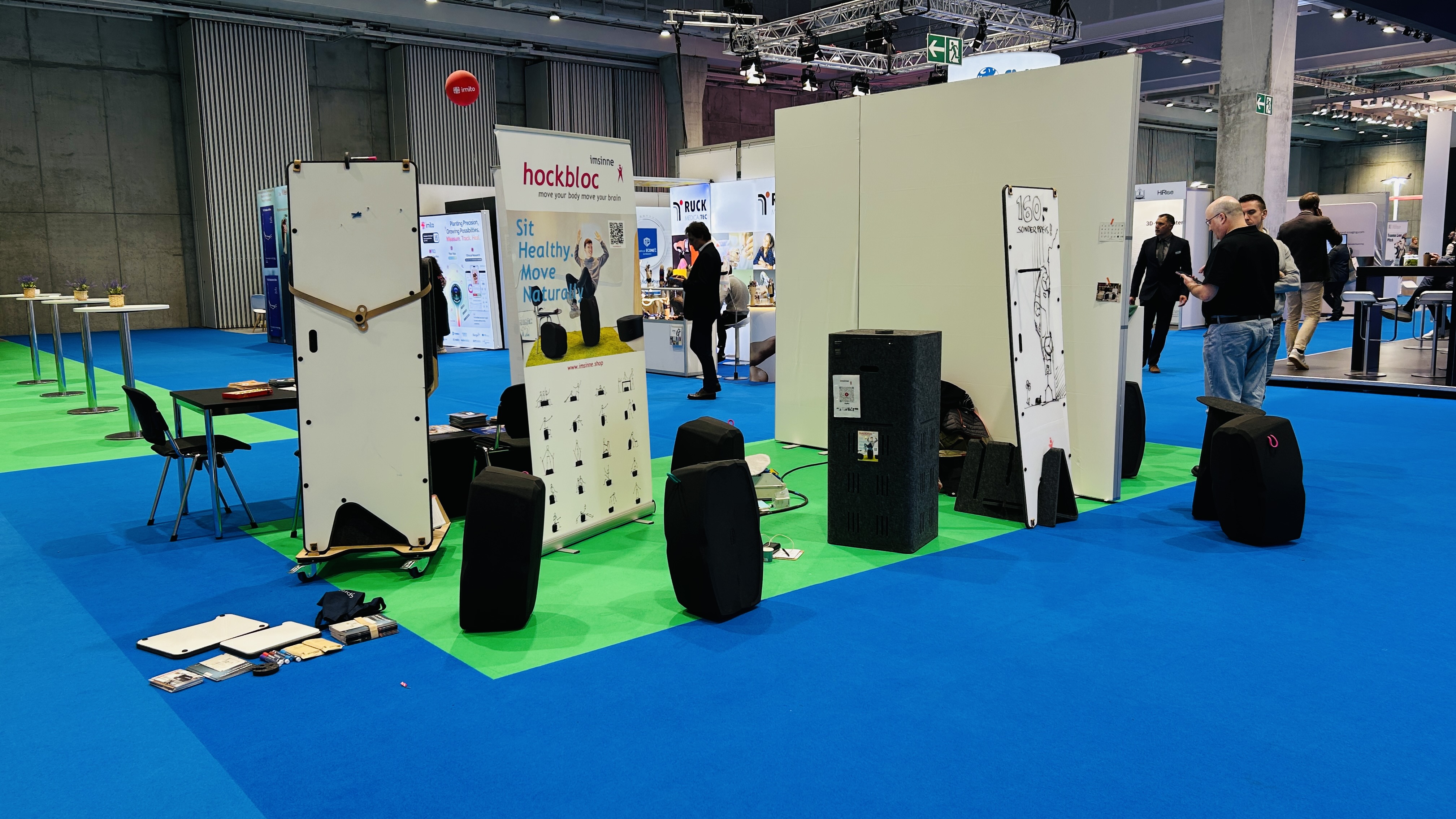 Ein heller Messestand bei der DKOU-Konferenz in Berlin. Im Vordergrund sind auf blauem und grünem Teppich verschiedene Produkte von "hockbloc" und "imsinne" zu sehen, darunter schwarze, organisch geformte Hocker und vertikale Präsentationswände mit Produktinformationen und Grafiken. Im Hintergrund sind weitere Stände und einige Konferenzbesucher zu erkennen, die sich umsehen oder miteinander sprechen. Ein großer roter Ball schwebt nahe der Decke. Der Stand wirkt offen und einladend.