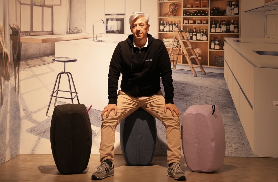 Tobias Greissing sitzt auf einem blauen hockbloc zwischen einem schwarzen und einem lavendelfarbenen hockbloc in einer modern gestalteten Küchenumgebung.