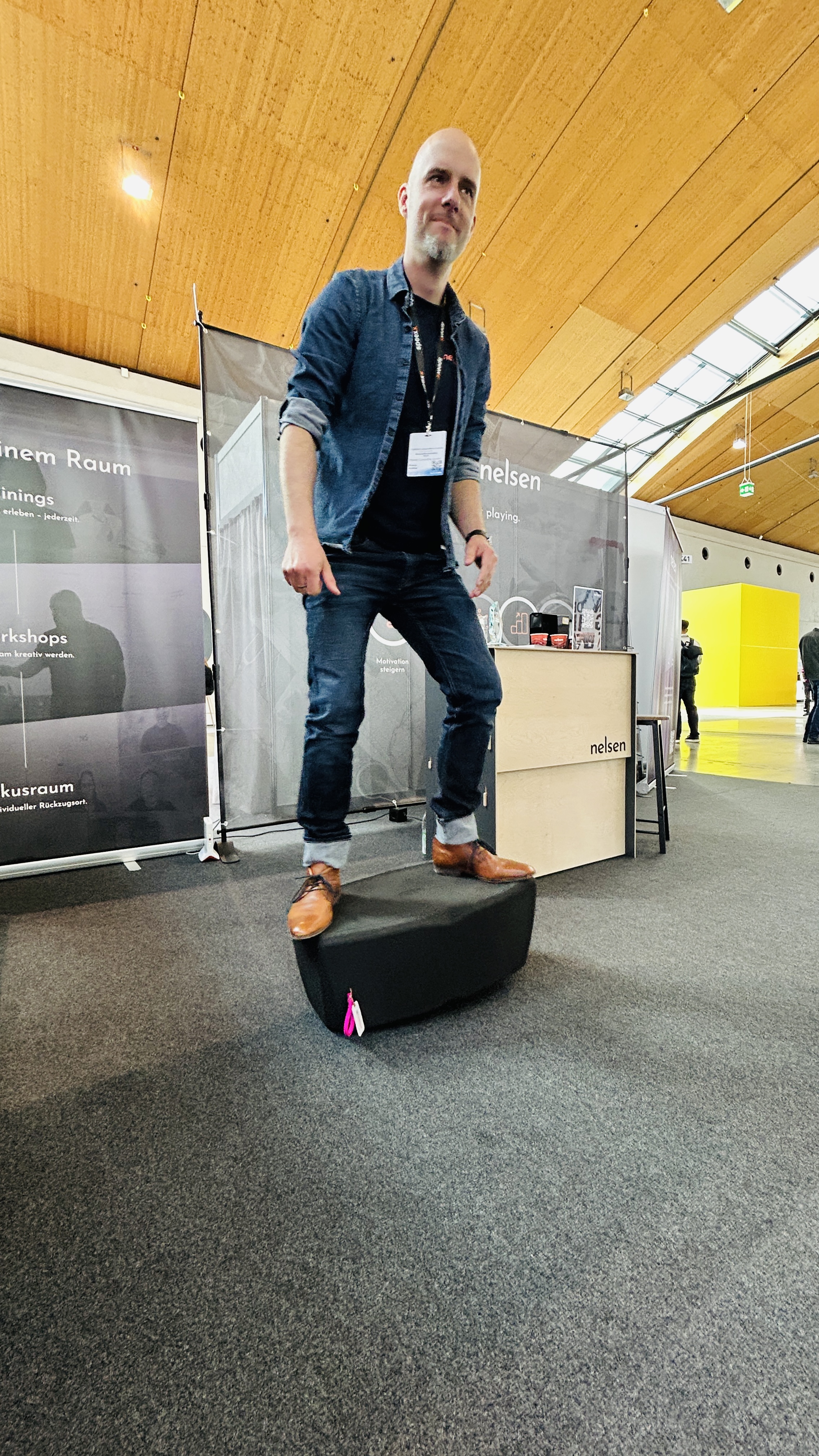 Ein lächelnder Mann in Denim-Kleidung und braunen Lederschuhen steht auf einem schwarzen, ergonomischen Sitzmöbel (wahrscheinlich ein Nelsen Hocker) an einem Messestand. Der Hintergrund zeigt einen geräumigen Messestand mit Werbebannern für "Nelsen" und "Workshops", einem Pult und anderen Besuchern in einer modernen Messehalle mit Holztäfelung und Dachfenstern.