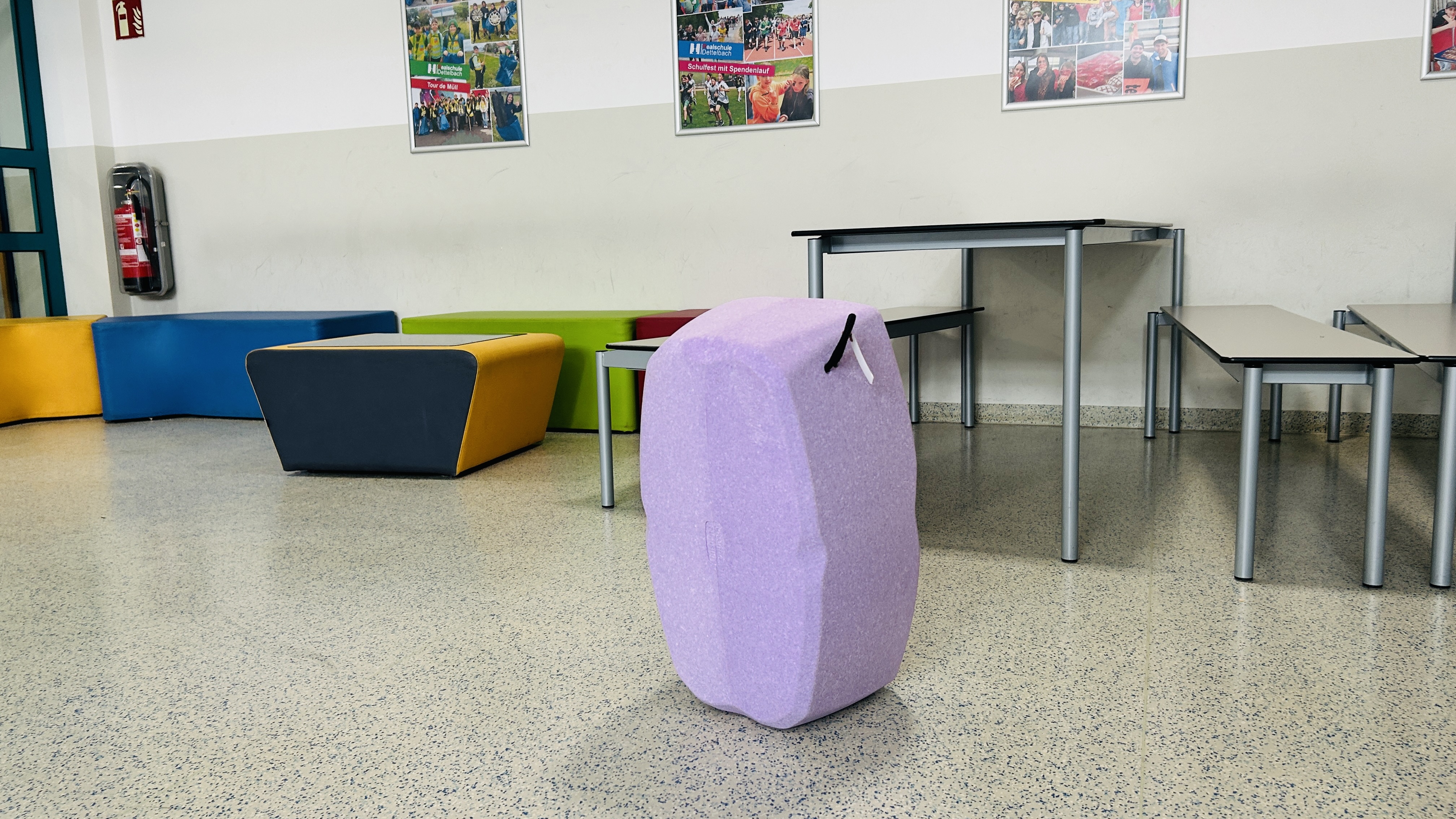 Ein lila, ergonomischer hockblox Sitzbaustein steht hochkant im Vordergrund eines hellen Schulflurs. Im Hintergrund befinden sich weitere bunte Polstermöbel in Gelb, Blau und Grün sowie moderne Tischgruppen mit Bänken für flexibles Arbeiten nach dem Churer Modell.
