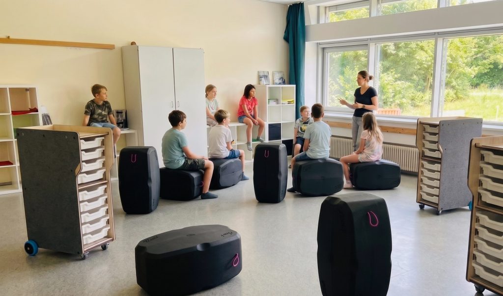Ein heller, einladender Klassenraum mit einer Grundschulklasse und einer Lehrerin im Casual-Look vor einer großen Fensterfront. Die Kinder sitzen flexibel im Halbkreis auf innovativen, dunkelgrauen Polsterhockern in Kofferform oder auf einer weißen Regalbank im Hintergrund. Im Raum verteilt stehen mobile, holzfarbene Rollcontainer mit weißen Materialeinschüben auf blauen Rollen. Die Atmosphäre ist unbeschwert, freundlich und zeigt ein klassisches, aber modernisiertes Grundschulszenario.