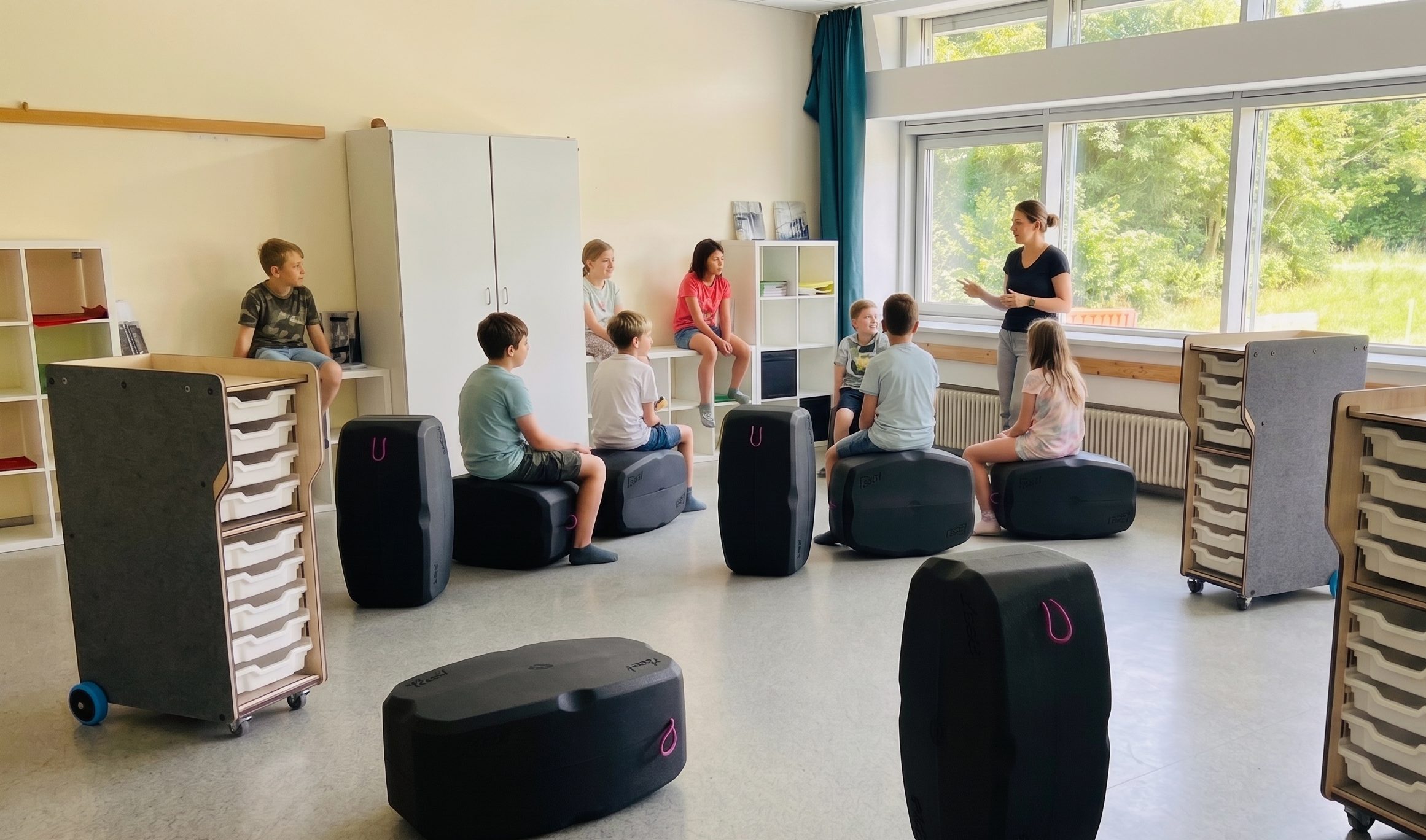 Ein heller, einladender Klassenraum mit einer Grundschulklasse und einer Lehrerin im Casual-Look vor einer großen Fensterfront. Die Kinder sitzen flexibel im Halbkreis auf innovativen, dunkelgrauen Polsterhockern in Kofferform oder auf einer weißen Regalbank im Hintergrund. Im Raum verteilt stehen mobile, holzfarbene Rollcontainer mit weißen Materialeinschüben auf blauen Rollen. Die Atmosphäre ist unbeschwert, freundlich und zeigt ein klassisches, aber modernisiertes Grundschulszenario.