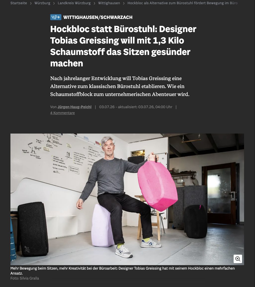 Ein Mann sitzt dynamisch auf einem ergonomischen, hellvioletten Schaumstoffhocker namens „Hockbloc“ in einer Werkstatt oder einem Büro. In der linken Hand hält er einen weiteren, auffallend pinkfarbenen Hockbloc-Hocker in die Höhe, um dessen Leichtigkeit zu demonstrieren. Er trägt ein graues Langarmshirt, dunkle Hosen und Sneaker. Im Hintergrund ist eine weiße Tafel mit Skizzen sowie eine Werkstattumgebung zu sehen. Das Bild stammt aus einem Artikel der Main-Post über den Designer Tobias Greissing.